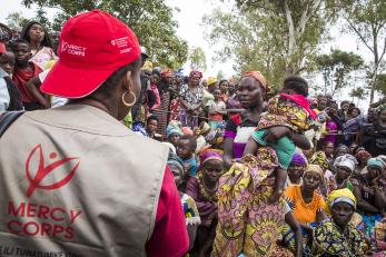A woman in a mercy corps hat and vest addresses a crowd of people gathered in dr congo